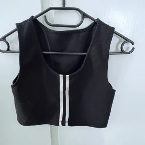 Svart croppad linnetopp med vita ränder - Snygg svart croppad linnetopp med två vita vertikala ränder framtill. Toppen är ärmlös och har rund halsringning. Perfekt för en trendig och sportig look.