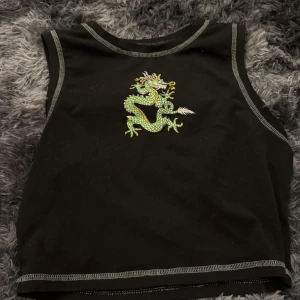 Svart croppad linnetopp med drakmotiv - Svart croppad linnetopp med grönt och gult drakmotiv på bröstet. Toppens kanter har kontrastsömmar i grått. Perfekt för en edgy och trendig stil. Storlek S