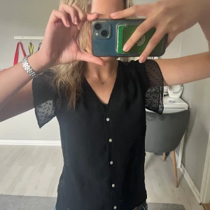 Svart blus med prickigt mönster från H&M - Säljer en svart blus från H&M med korta, transparenta ärmar och diskret prickigt mönster. Blusen har v-ringning och knappar framtill.