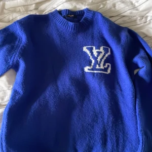 Blå stickad tröja med vit LV-logga - Säljer en blå stickad tröja från Louis Vuitton med vit LV-logga på bröstet. Tröjan har rund halsringning och långa ärmar med ribbade muddar. Perfekt för dig som gillar klassiska märken och vill ha en snygg statement-piece.