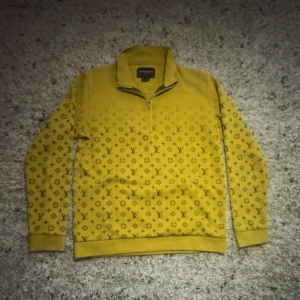 Gul Louis Vuitton ziptröja - Gul långärmad tröja från Louis Vuitton med halv dragkedja och klassiskt LV-mönster över hela plagget. Tröjan har ståkrage och ribbade muddar vid ärmslut och nederkant. Perfekt för dig som vill sticka ut med en exklusiv look.
