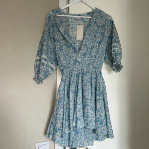 Blå blommig klänning - Supersöt blå och vit kortklänning köpt utomlands. Klänningen har v-ringning och puffiga kortärmar. Perfekt för sommarens alla tillfällen! Aldrig använd, prislapp kvar!