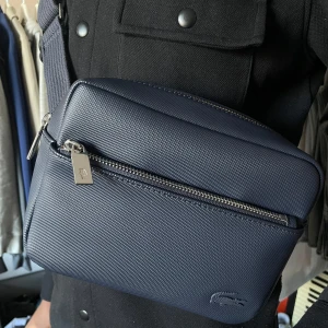 Mörkblå axelväska från Lacoste - Snygg mörkblå axelväska från Lacoste med strukturmönstrad yta och silverfärgade dragkedjor. Väskan har ett diskret Lacoste-logotyp framtill och justerbar axelrem. Perfekt för att bära det viktigaste med stil.🙌🏾Lägg gärna egna prisförslag!! Säljer då min bror inte använder den, precis som ny🤩‼️