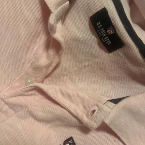 Ljusrosa pikétröja från U.S. Polo Assn. - Säljer en ljusrosa pikétröja från U.S. Polo Assn. med klassisk krage och knäppning framtill. Tröjan har korta ärmar och en liten broderad logga på bröstet. Perfekt för en sportig och stilren look.