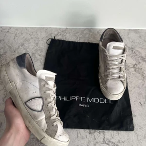 Vita sneakers från Philippe Model - Säljer ett par vita sneakers från Philippe Model med grå och svarta detaljer. Skorna har snörning, rund tå och platt sula. Klassisk design med logotyp på sidan och ovandel i skinn och mocka.