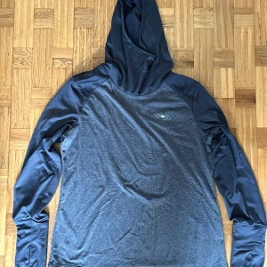 Mörkgrå hoodie från Crivit i storlek L - Sportig mörkgrå hoodie från Crivit med släta svarta ärmar och huva. Tröjan har lång ärm och är tillverkad i ett mjukt, syntetiskt material som passar perfekt till träning eller chill. Liten logga på bröstet och tumhål vid ärmslut.
