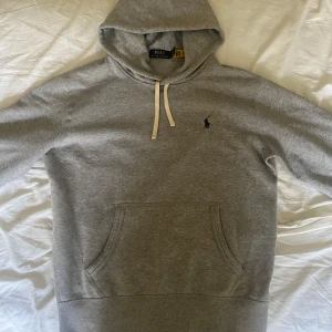 Grå hoodie från Polo Ralph Lauren - Klassisk grå hoodie från Polo Ralph Lauren med broderad logga på bröstet. Tröjan har huva med vita snören, känguruficka och långa ärmar. Perfekt för chill dagar och enkel att matcha med jeans eller joggers. Helt ny och aldrig använd strlk S.