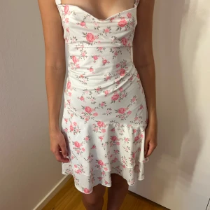 Vit blommig kortklänning med volang - Supersöt vit kortklänning med rosa blommigt mönster och volang nertill. Klänningen har smala axelband, lätt draperad byst och är figurnära med en flowy kjol. Perfekt för sommaren.