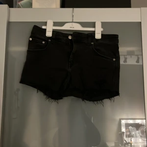 Svarta jeansshorts från 157, M - Svarta jeansshorts från 157 med råa, fransiga kanter och klassiska fem fickor. Modellen är midwaist och har silverfärgade knappar och nitar. Perfekta för en chill sommarlook med lite edge.