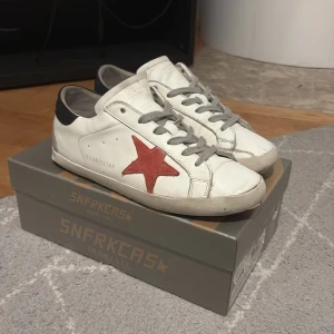 Golden Goose vita sneakers med röd stjärna - Golden Goose sneakers i vitt skinn med ikonisk röd stjärna på sidan och grå snörning. Skorna har svart detalj på hälen och en platt, ljusbrun sula. Perfekt för dig som gillar en cool och avslappnad streetstil. De är lite slitna i hälen på under sidan av skon anars är de rätt så fina.