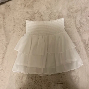 Vit volangkjol från Chlesa - Supersöt vit kortkjol från Chlesa med bred smock i midjan och dubbla lager volanger. Kjolen är luftig och har en flowy känsla, perfekt för varma dagar. Tillverkad i ett tunt, lätt material som ger en romantisk vibe.