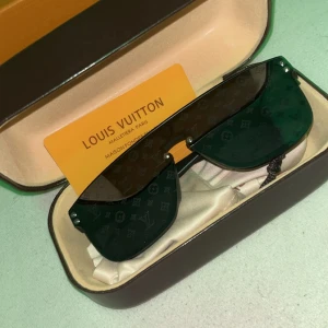 Svarta Louis Vuitton solglasögon - Säljer ett par svarta Louis Vuitton solglasögon med breda bågar och shield-formade glas. Glasen har diskret LV-monogram tryckt över hela ytan för en lyxig vibe. Snygg och modern design, perfekt för dig som vill sticka ut med exklusiva accessoarer.