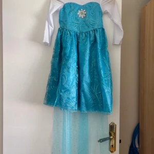 Blå prinsessklänning med glitterdetaljer - Superfin blå prinsessklänning med långa vita ärmar, glittrigt mönster och en stor silverfärgad snöflinga framtill. Klänningen har en lång, transparent släp med glitterprickar och vackra broderade detaljer. Perfekt för dig som älskar frostiga sagor!