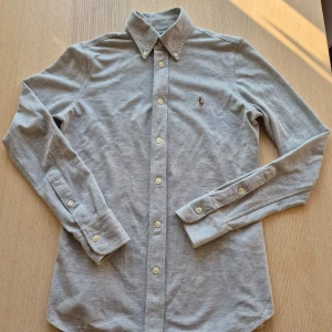 Grå skjorta från Ralph Lauren - Snygg grå skjorta i nyskick från Ralph Lauren med klassisk button-down krage och broderad logga på bröstet. Skjortan är långärmad och har vita knappar framtill och vid ärmsluten. Tillverkad i mjuk bomull för en bekväm känsla.