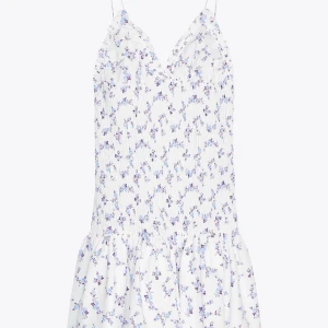 Vit blommig kortklänning med smock - Supersöt vit kortklänning med små blå och lila blommor, smockad överdel och tunna axelband. Klänningen har v-ringning och volangkant nertill, perfekt för varma dagar. Lätt och luftig känsla, passar dig som gillar romantisk och somrig stil.