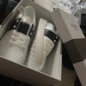 Valentino Garavani Open sneakers vit/svart - Snygga Valentino Garavani Open sneakers i vitt skinn med svart bred rem över mitten och coola nitar på sulan. Klassisk rund tå, vita snören och chunky platt sula med mönstrad undersida. Perfekta för dig som vill sticka ut med lyxig streetstyle.