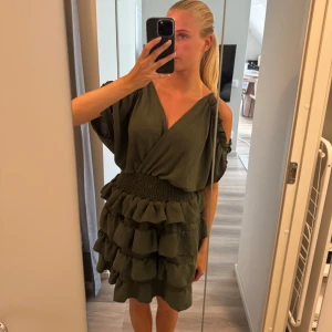 Olivgrön volangklänning omlott - Supersöt olivgrön cocktailklänning med omlottliv, djup v-ringning och bred smock i midjan. Oanvänd, Kommer från märket Wal.G, nypris 529kr