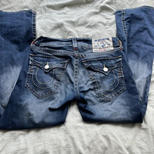 True Religion bootcut jeans blå - Säljer ett par sjukt snygga blå bootcut jeans från True Religion med snygga kontrastsömmar och ikoniska fickdetaljer bak. Jeansen har klassisk femficksdesign, markerade nitar och en cool tvättad look.💕