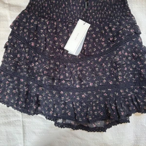 Blommig volangkjol från Neo Noir - Supersöt svart kortkjol från Neo Noir i storlek 34 med små blommor i rosa och grönt. Kjolen har flera volanger, smock i midjan och spetsdetaljer längs kanterna. Helt ny med prislappen kvar💕