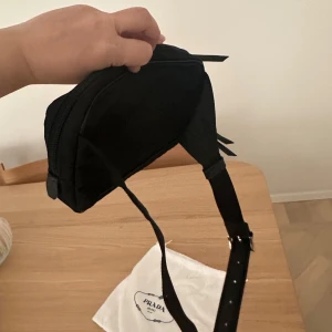 Svart axelväska från Prada i nylon - Snygg svart axelväska från Prada i klassisk rektangulär form. Väskan är gjord i slitstark nylon med justerbar axelrem och har Pradas ikoniska metallogo framtill. Praktisk dragkedja upptill och extra fack på framsidan för småsaker.