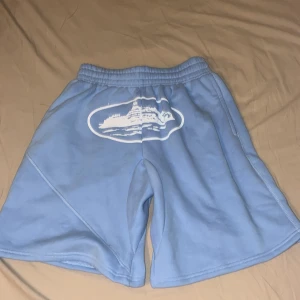 Blå fleece shorts från Corteiz - Ljusblåa fleece shorts från Corteiz med elastisk midja och tryckt vit grafik framtill. Modellen har en loose passform, sidofickor och en bakficka. Perfekta för chill dagar eller när du vill ha något bekvämt och snyggt på sommaren.