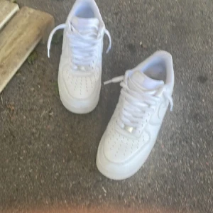 Vita Nike Air Force 1 sneakers - Klassiska helvita Nike Air Force 1 sneakers med snörning och perforerade detaljer på tån. Skorna har en platt sula och är tillverkade i skinn, vilket ger en clean och tidlös look som alltid funkar.