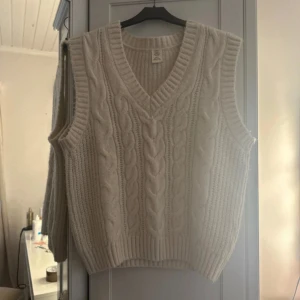 Beige stickad väst med kabelmönster - Beige stickad väst från urban outfitters  med grovt kabelstickat mönster framtill och ribbade kanter. Västen har v-ringning och en loose passform, perfekt att slänga över en skjorta eller t-shirt för en chill look.