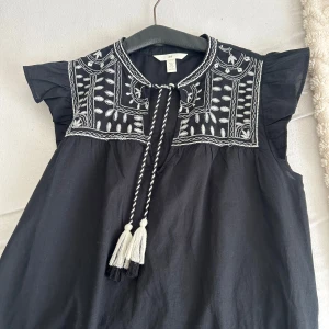 Svart broderad blus H&M S - Svart blus från H&M med vit brodyr upptill och snygga volangärmar. Blusen har knytband med tofsar vid halsen och ett luftigt, rakt fall. Perfekt för dig som gillar boho-vibbar och detaljerad design. Storlek S men passar mig som är XXS-XS, om du gillar det stort, använd få gånger. Nypris 260-280kr