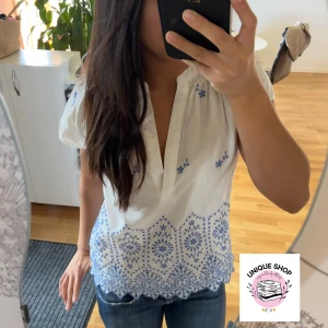 Vit blus med blå broderi - Supersöt vit blus med blått broderat mönster och v-ringning. Blusen har korta ärmar och ett detaljerat broderi längs nederkanten och små blommor upptill. Perfekt för en fräsch och somrig look. Materialet känns lätt och luftigt.