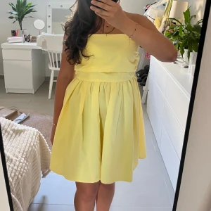 Gul axelbandslös kortklänning - Superfin gul axelbandslös kortklänning med voluminös kjol och smockad rygg. Perfekt för dig som vill ha en söt och somrig look. Klänningen har en enkel, stilren design och är lätt att matcha med accessoarer. Storlek M men passar oxå S och L eftersom den har resor på baksidan. Använd en gång.
