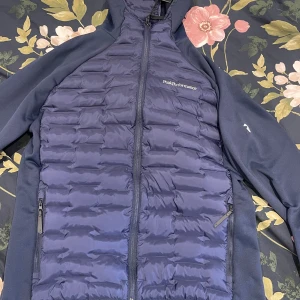 Peak Performance jacka - Snygg marinblå pufferjacka från Peak Performance med quiltad framsida och rygg, huva och dragkedja. Jackan har två fickor med dragkedja och logga på bröstet. Perfekt för kyliga dagar när du vill hålla stilen.