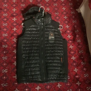 Svart dunväst Geographical Norway S - Svart quiltad dunväst från Geographical Norway, modell GNX 850. Västen har huva, dragkedja framtill och två fickor med dragkedja. Snygg badge på bröstet och orange detaljer. Perfekt för lager-på-lager och streetstyle.