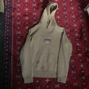 Beige hoodie från GANT med logga - Snygg beige hoodie från GANT med broderad logga och amerikanskinspirerat emblem på bröstet. Klassisk känguruficka framtill och huva. Perfekt för dig som gillar stilren och avslappnad look.