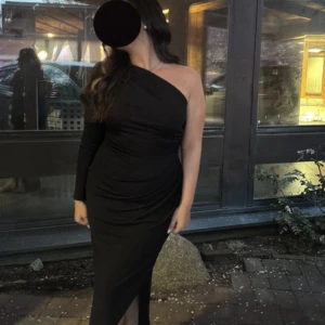 Svart långklänning one shoulder - Säljer en elegant svart långklänning med oneshoulder-design och lång ärm på ena sidan. Klänningen har figurnära passform och en snygg slits framtill. Perfekt för dig som vill sticka ut med en stilren look.