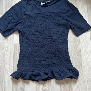 Svart smockad topp med volang Gina Tricot - Svart topp från Gina Tricot med smockad kropp och volangkant nertill. Toppen har korta ärmar och är gjord i mjuk bomull. Perfekt för dig som gillar en söt och trendig stil med extra detaljer.