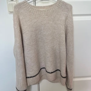 Beige stickad tröja från Lindex - Beige stickad tröja från Lindex med svarta broderade detaljer längs ärmar och nederkant. Tröjan har rund halsringning, lång ärm och en loose fit. Inte använd så många gånger. Ganska lång. 