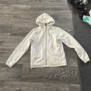 Vit Sail Racing hoodiejacka 160 - Vit hoodiejacka från Sail Racing med dragkedja framtill och broderad logga på bröstet och huvan. Jackan har två fickor med dragkedja och är tillverkad i mjukt material som känns skönt mot huden. Perfekt för dig som gillar stilren och sportig look.