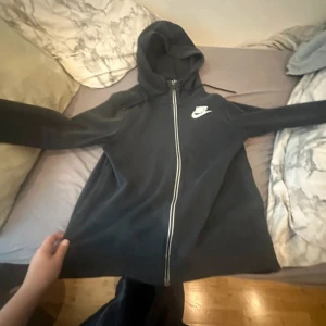 Svart zip-hoodie från Nike, stl S - Svart hoodie från Nike med dragkedja hela vägen och vit Nike-logga på bröstet. Klassisk modell med huva och snörning, tillverkad i mjukt bomullsmaterial. Perfekt för chill dagar eller när du vill ha en sportig look.