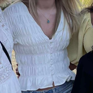 Vit blus med volang och knappar - Supersöt vit blus med v-ringning från gina, volangkant nertill och smockad midja. Blusen har långa ärmar och knappar framtill. 