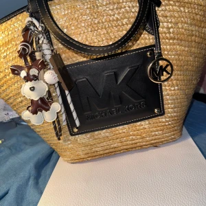 Stråväska från Michael Kors med svarta detaljer - Snygg shoppingväska från Michael Kors i flätad strådesign med svarta skinnhandtag och stor svart logotyp på framsidan. Väskan har en praktisk snörning upptill och dekorativa nyckelringar. Perfekt för dig som vill ha en trendig och rymlig väska till sommaren.