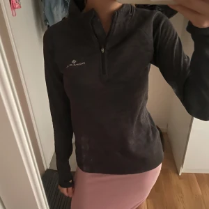 Svart långärmad träningströja Ronhill - Svart långärmad träningströja från Ronhill med halv dragkedja framtill och diskret logga på bröstet. Tröjan har en sportig passform och är tillverkad i ett lätt syntetmaterial som andas, perfekt för löpning eller gym.