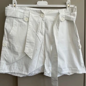Vita  shorts med knytband - Snygga vita shorts med hög midja och bred linning, dekorerade med stora vita knappar och ett knytband i midjan. Shortsen har fickor fram och bak samt detaljerade sömmar. Perfekta för sommarens alla äventyr.