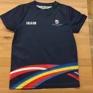Lilla EM Handbolls-EM 2020 t-shirt - Mörkblå t-shirt från Lilla EM, officiell merchandise för Handbolls-EM 2020 i Malmö. Tryck med färgglada vågor i rött, blått, gult och vitt nedtill samt loggor och text fram och bak. Tillverkad i lätt och sportig polyester, perfekt för handboll eller träning.