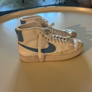 Nike Blazer Mid vit/blå sneakers - Snygga Nike Blazer Mid sneakers i vitt skinn med ljusblå swoosh och hälpanel. Klassisk retrostil med högt skaft, snörning och platt sula. Perfekta för dig som gillar streetwear och vill sticka ut med en fräsch färgkombo.