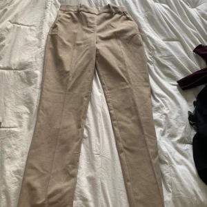 Beige kostymbyxor i storlek 34 - Snygga beige kostymbyxor med raka ben och pressveck. Byxorna har hällor för bälte och klassisk passform. Perfekta för en clean och stilren look. Materialet känns som bomullsmix och färgen är neutral beige.