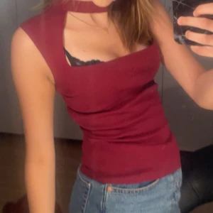 Vinröd ärmlös topp med choker-detalj - Säljer en vinröd ärmlös topp med unik choker-detalj runt halsen och breda axelband. Toppen har en tight passform och en snygg, rak skärning vid bysten. Perfekt att styla med jeans eller kjol för en trendig look.