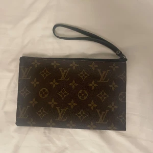 monogram kuvertväska - Snygg kuvertväska med klassiskt monogrammönster i brunt och beige. Väskan har en handledsrem i svart skinn och stängs med dragkedja. Perfekt storlek för det viktigaste och en riktig statement-accessoar. Aldrig använd är 21x14 cm.