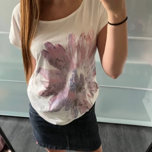 Vit t-shirt med stor blommig print - Vit t-shirt med korta ärmar och en stor, färgglad blomma i rosa och lila toner framtill. T-shirten har en avslappnad passform och är gjord i mjuk bomull, perfekt för en chill och snygg look. Rund halsringning och enkel baksida utan tryck.