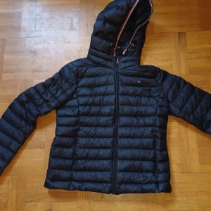 Svart dunjacka från Tommy Hilfiger - Snygg svart dunjacka från Tommy Hilfiger med huva och dragkedja framtill. Jackan har klassiska quiltade paneler och en liten logga på bröstet. Perfekt för kyliga dagar och har en sportig vibe med stilrena detaljer. Hör av er med frågor eller snabb affär. Pris kan diskuteras
