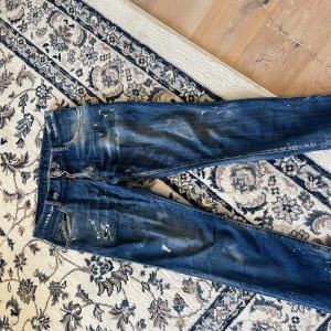Dsquared2 blå jeans med slitningar - Snygga blå jeans från Dsquared2 med coola slitningar och färgstänk för en edgy look. Klassisk femficksmodell med röd Dsquared2-tag vid gylfen och läderpatch bak. Jeansen har en rak passform och är tillverkade i bomull.ÄKTA KÖPTES FÖR 5400 av dsq 